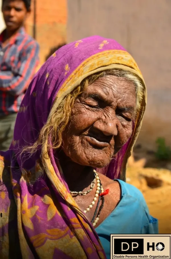 A deeply wrinkled elderly woman with eyes closed in reflection and a serene expression, wearing a purple and gold head covering and necklaces. The Disable Persons Health Organization (DP-HO) logo is in the bottom right corner, suggesting support and community to counteract potential Social Isolation in older age.