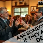 A medium shot photograph featuring two middle-aged men in suits facing each other across a wooden table in a busy, warm pub setting. The man on the left, with grey hair and beard, and the man on the right, with dark hair and glasses, are actively signing, their open hands held towards each other in gesture. Background people are softly blurred, indicating other patrons and decor. The atmosphere is respectful and engaged. In the upper-right corner is the complete black and white DP HO Disable Persons Health Organization website logo. Overlaying the bottom-right corner is a large, translucent, diagonal white text band containing the bold, black text 'ASK SOMEONE IF THEY ARE DEAF', with a faint diamond-shaped accent. The focus is sharp on the two main subjects, conveying connection and respectful communication etiquette."