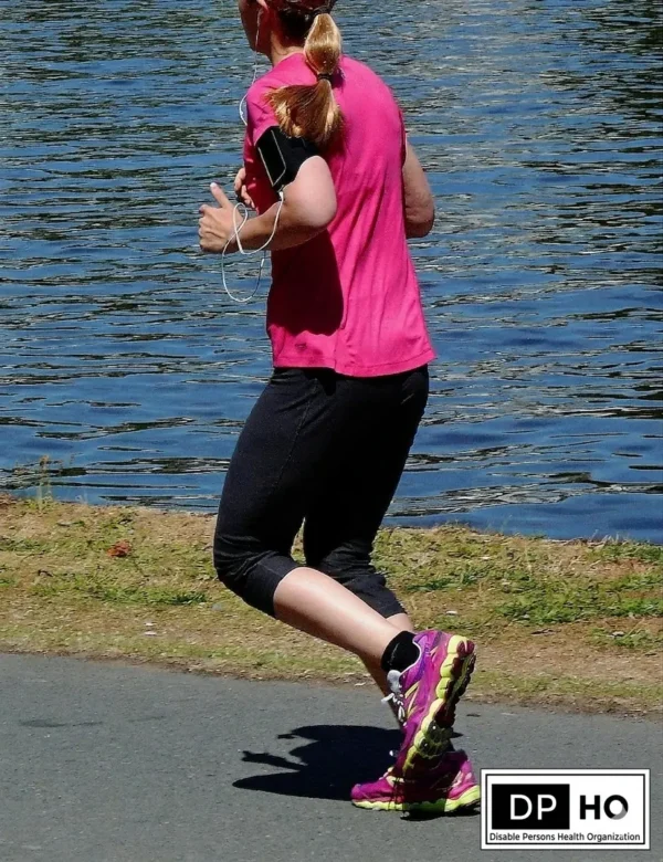 A woman in a pink shirt and black shorts running along the side of a body of water, wearing a waist pack and carrying a D P H O water bottle with the logo visible.