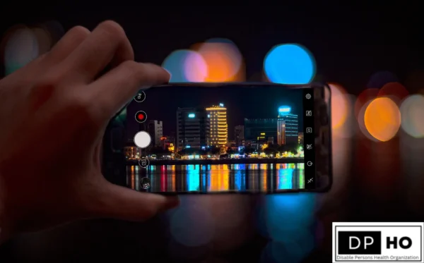A first-person view of a hand holding a smartphone, capturing a crisp, clear photo of a brightly lit city skyline at night reflected over water. The background is a soft bokeh of colorful, out-of-focus lights, contrasting with the sharp digital image on the screen. This serves as a metaphor for how AI and Speech-to-Text tools act as a lens, helping to clarify communication and explain why deaf people often not type things normally by bridging the gap between different linguistic worlds. The DP-HO (Disable Persons Health Organization) logo is featured in the bottom right corner.