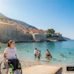 A bright, sunny photograph of a coastal beach in Croatia, featuring a young woman in a green and black wheelchair smiling on a wooden accessible ramp. In the background, people are swimming in clear turquoise water against the backdrop of a historic Mediterranean walled city and hills. A blue sign with a wheelchair icon and Croatian text is visible in the foreground, and the "Disable Persons Health Organization" (DP HO) logo is placed in the bottom right corner.