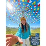 A smiling female hiker in a blue jacket and yellow beanie takes a selfie in a scenic mountain valley under a bright, sunny sky. Colorful prayer flags spiral in a large canopy overhead, and small white petals drift through the air. In the bottom right corner, a blue and white text block features the International Symbol of Access (wheelchair icon) and reads: "11 TYPES OF MOBILITY DISABILITIES: THE 2026-27 GUIDE." A "Special Ed Expert Ali" YouTube channel logo is in the top-left corner, and a "DP HO - Disable Persons Health Organization" logo is in the bottom-right corner.