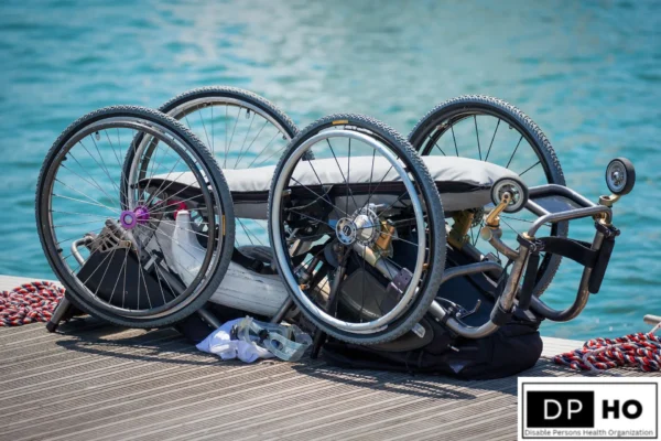 A high-quality photo of two specialized athletic wheelchairs resting on a wooden deck by the water. The wheelchairs feature lightweight frames, large spoked wheels with purple hubs, and specialized padding for high-performance use. A white towel and swimming goggles lie nearby, symbolizing the intersection of sport and accessibility. The Disable Persons Health Organization (DP-HO) logo is positioned in the bottom right corner.